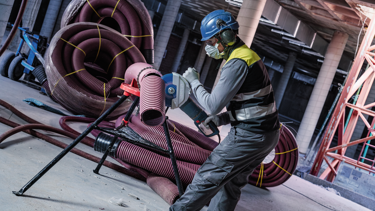 A person wearing safety equipment is cutting a plastic pipe in a construction area.