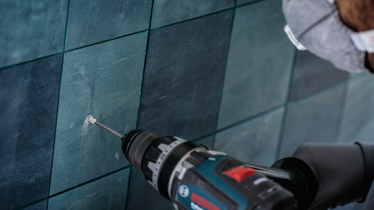 Person wearing safety equipment drills a hole into a tiled wall.