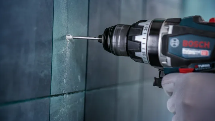 Person wearing safety equipment drills into a tiled wall.