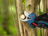 A person wearing safety equipment uses a cordless pruner to cut a tree branch.