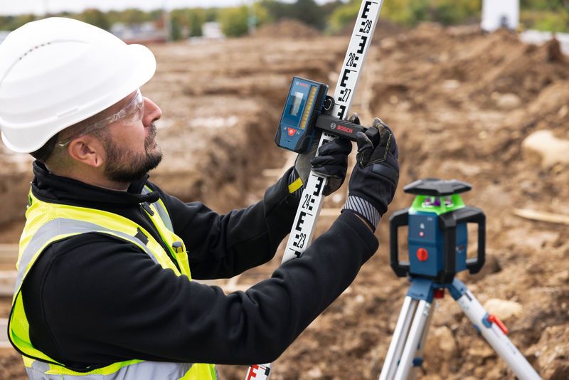 A person wearing safety equipment adjusts a laser receiver on a leveling rod near a laser leveling tool.