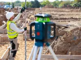 A person wearing safety equipment uses a laser leveling tool and receiver at a construction site.