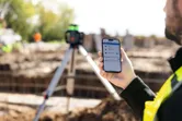 A person wearing safety equipment uses a smartphone to control a laser leveling tool outdoors.