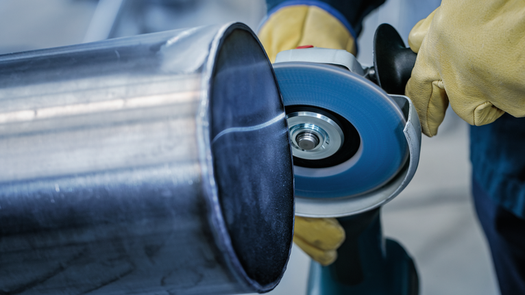 Person wearing safety equipment cuts a metal pipe with an angle grinder.