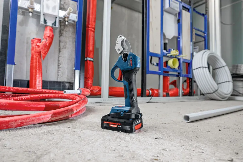Cordless pipe shear stands on a construction floor surrounded by pipes and plumbing materials.