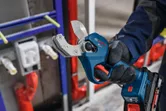 A person wearing safety equipment holds a cordless pipe shear near plumbing pipes.