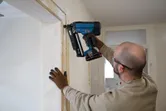 A person wearing safety equipment uses a cordless finish nailer on a door frame.