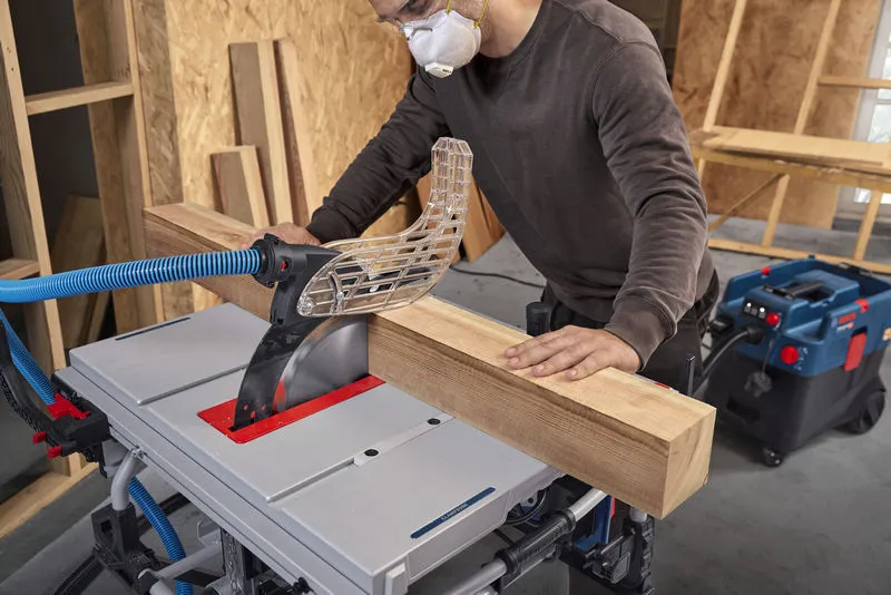 A person wearing safety equipment cuts a thick wooden beam using a table saw.