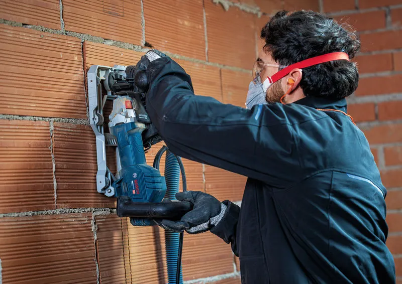 A person wearing safety equipment uses a wall chaser to cut grooves in a brick wall.