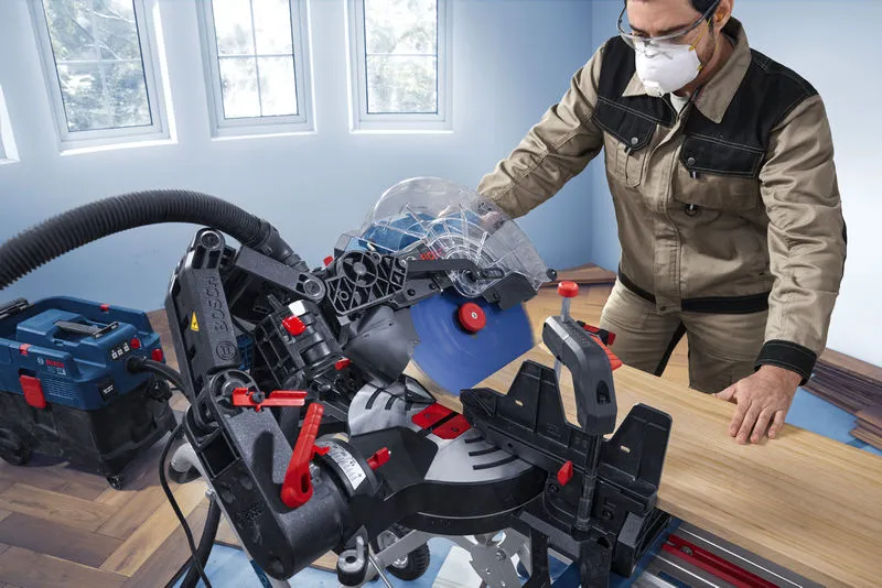 A person wearing safety equipment operates a mitre saw to cut a wooden plank.