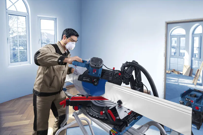 A person wearing safety equipment operates a mitre saw to cut trim in a well-lit room.