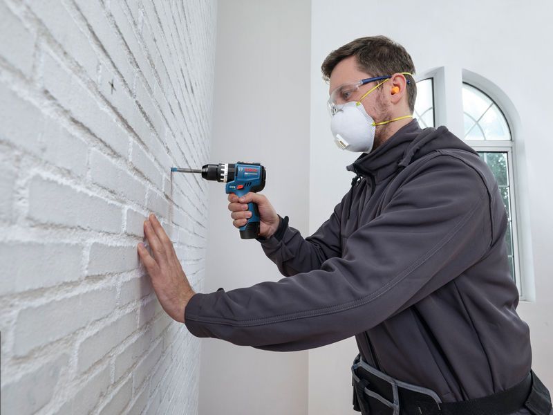 A person wearing safety equipment drills into a white brick wall.
