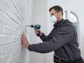 A person wearing safety equipment drills into a white brick wall.