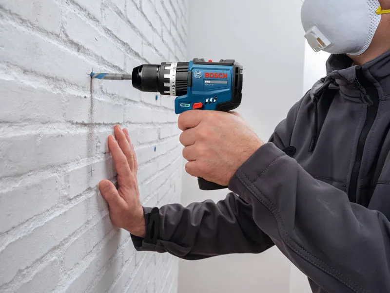 A person wearing safety equipment drills into a brick wall with a cordless impact drill.