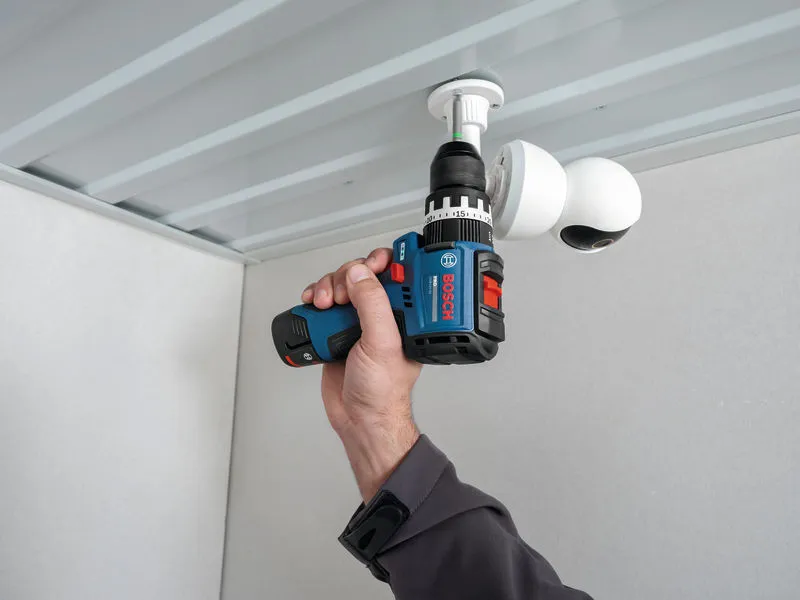A person uses a cordless impact drill driver to install a surveillance camera.