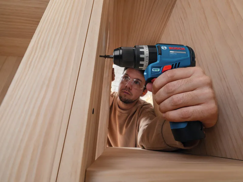 A person wearing safety equipment uses a cordless impact drill on a wooden cabinet.