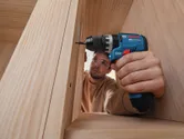 A person wearing safety equipment uses a cordless impact drill on a wooden cabinet.