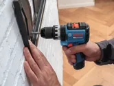 A person uses a cordless impact drill to install a bracket on a white brick wall.