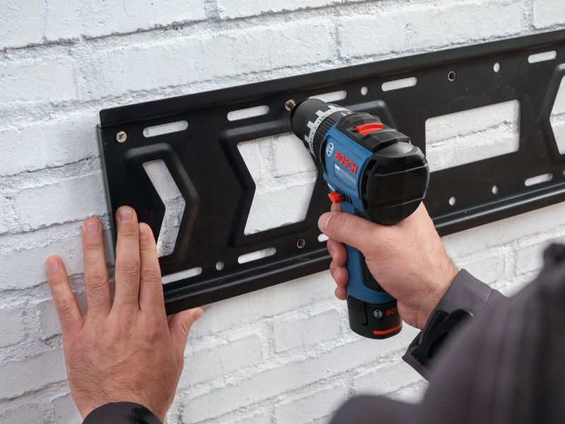 A person mounts a metal bracket on a white brick wall using a cordless impact drill.