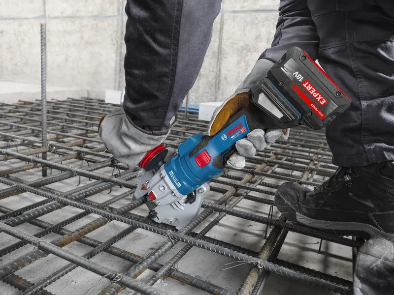 A person wearing safety equipment uses a cordless rebar cutter on a steel grid.