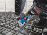 A person wearing safety equipment uses a cordless rebar cutter on a steel grid.