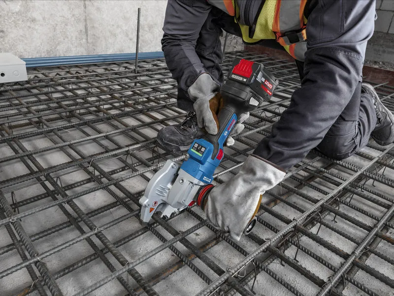 A person wearing safety equipment uses a cordless tool to cut rebar on a construction site.