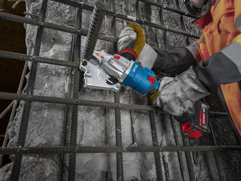 A person wearing safety equipment uses a cordless tool to cut through rebar on a concrete surface.