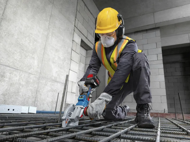 A worker wearing safety equipment uses a cordless rebar cutter on steel rods.
