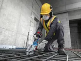 A worker wearing safety equipment uses a cordless rebar cutter on steel rods.