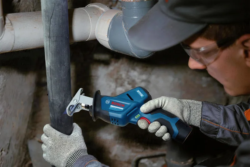 A person wearing safety equipment cuts a plastic pipe with a cordless saw.