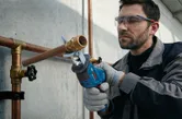 A person wearing safety equipment cuts a metal pipe with a power tool.