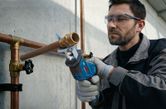 A person wearing safety equipment cuts a metal pipe with a power tool.