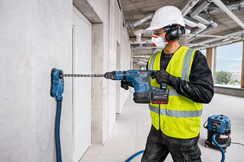 A person wearing safety equipment drills into concrete with a cordless rotary hammer.