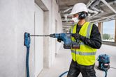 A person wearing safety equipment drills into concrete with a cordless rotary hammer.