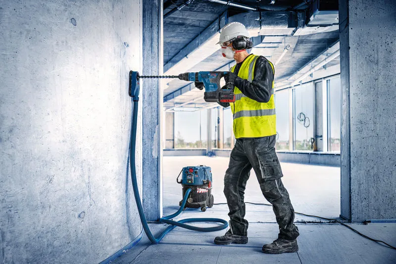 A worker wearing safety equipment drills into concrete with a cordless rotary hammer.