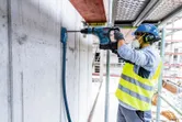 A person wearing safety equipment drills into a concrete wall with a cordless rotary hammer.