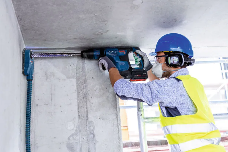 A person wearing safety equipment drills into a concrete ceiling with a cordless rotary hammer.