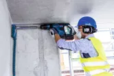 A person wearing safety equipment drills into a concrete ceiling with a cordless rotary hammer.
