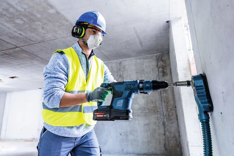 A person wearing safety equipment drills into concrete with a cordless rotary hammer.