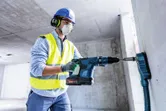 A person wearing safety equipment drills into concrete with a cordless rotary hammer.
