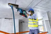 A person wearing safety equipment drills into a concrete ceiling with a rotary hammer.