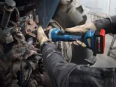 A person wearing safety equipment uses a cordless straight grinder on metal machinery.