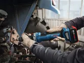 A person wearing safety equipment uses a cordless straight grinder on metal machinery.