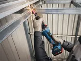 A person wearing safety equipment grinds a steel beam with a cordless straight grinder.