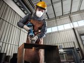 A person wearing safety equipment grinds metal with a cordless straight grinder.