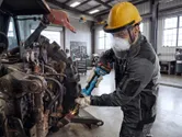 A person wearing safety equipment grinds metal on farm machinery in a workshop.