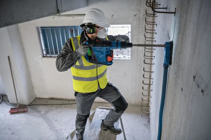 A person wearing safety equipment drills into a concrete wall with a rotary hammer.
