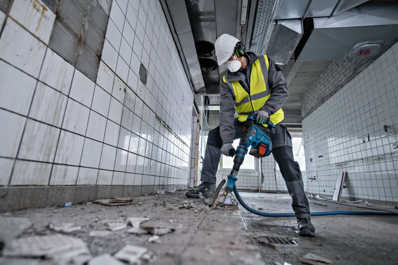 A person wearing safety equipment uses a rotary hammer to break tiled flooring.