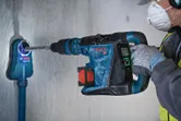 A person wearing safety equipment uses a rotary hammer to drill into a concrete wall.