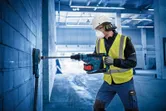 A person wearing safety equipment drills into a brick wall with a cordless rotary hammer.